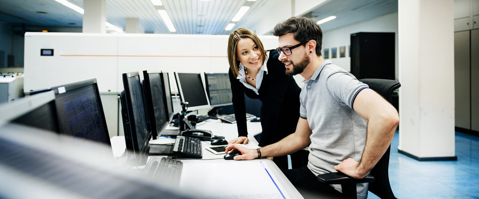 Eine Frau und ein Mann unterhalten sich, beide lächeln. Sie befinden sich in einem Großraumbüro mit vielen Computermonitoren.