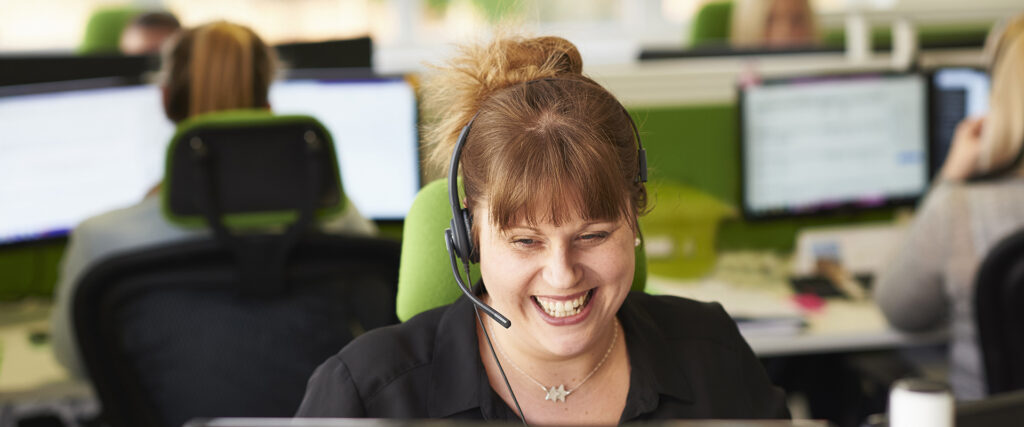 Eine Frau mit einem Headset lächelt. sie sitzt an einem PC-Arbeitsplatz.