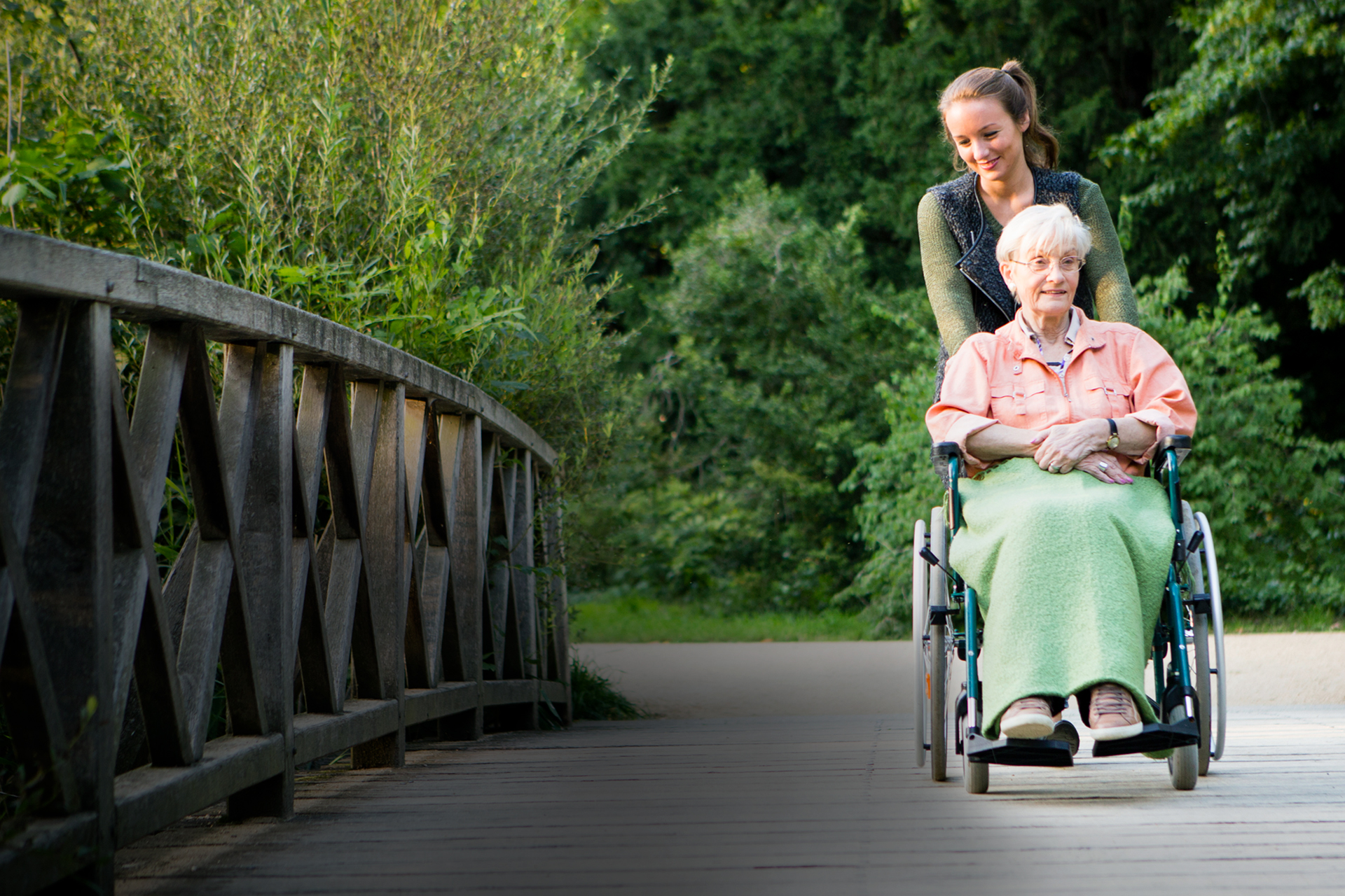 Eine junge Frau schiebt eine Seniorin im Rollstuhl über eine große Holzbrücke in einem Park.