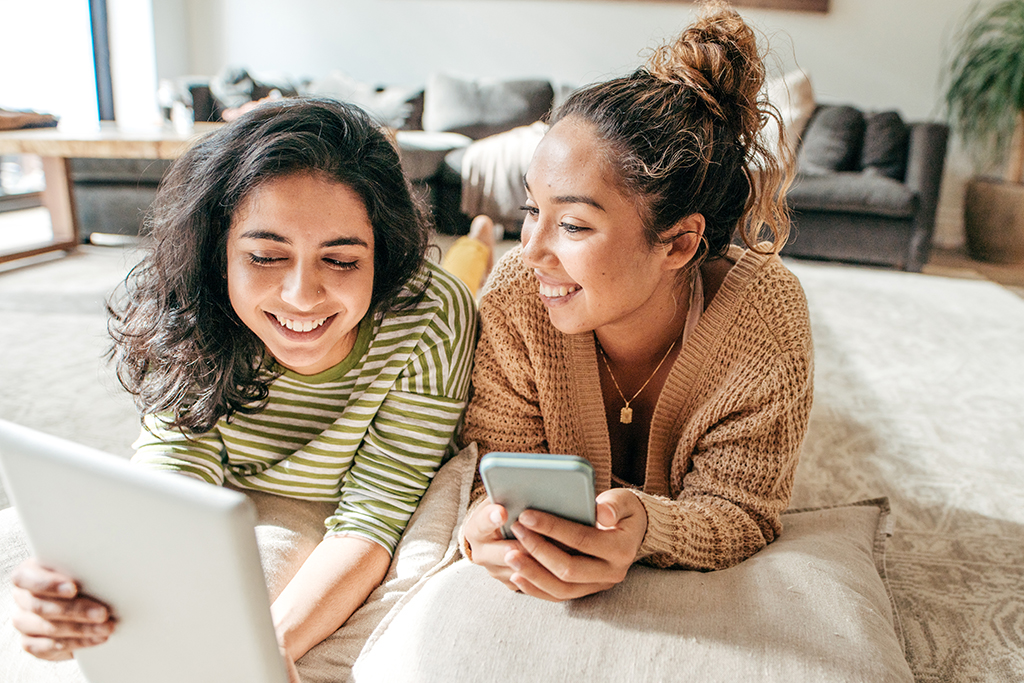 Zwei junge Frauen liegen bäuchlings auf einem Bett. Die eine hält ein Tablet in den Händen, die andere ein Smartphone. Beide lächeln.